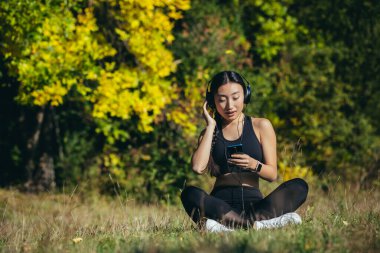 Genç Asyalı formda bir kadın Lotus pozisyonunda oturmuş meditasyon yapıyor ve açık havada müzik dinliyor. Mutlu dişi kız ormanda ya da parkta kulaklıkla doğanın tadını çıkarıyor. Sesle iyileştirme