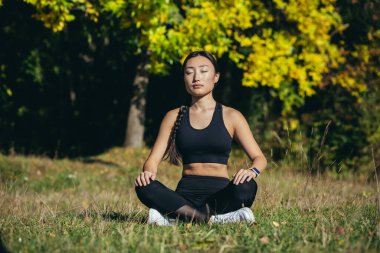 Young beautiful asian woman performs fitness yoga exercises, sitting in lotus position on the grass and meditating in the park