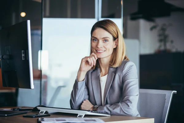 Happy and successful business woman boss working at computer in modern office looking at camera and smiling