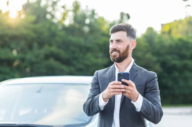 Neşeli ve başarılı erkek iş adamı araba satıcısı araba satmak için otoparkın yanındaki telefonu kullanıyor.