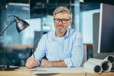 Deneyimli gri saçlı tasarımcı, gülümseyen ve kameraya bakan, yeni ofis merkezinin mimari projektörü üzerinde çalışan adam.