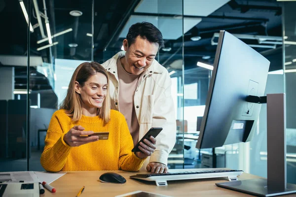 Girişimci Asyalı erkek ve kadınlar modern bir ofiste çalışıyorlar. Telefon ve banka kartlarıyla online alışveriş yapmaktan mutlular.