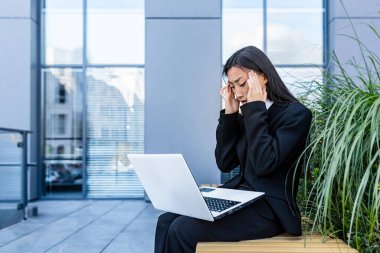 üzgün ve kafası karışmış Asyalı iş kadını laptopta çalışıyor, ofis dışında, bankta oturuyor.