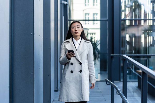 Beautiful Asian woman in business clothes uses the phone, walks near the modern office center outside
