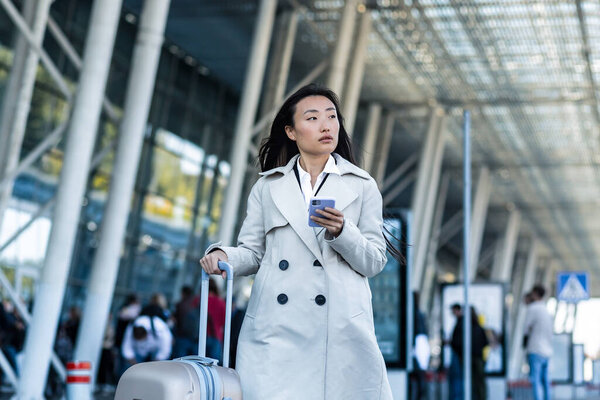 Beautiful Asian woman, near the airport uses a phone application for online booking, a tourist with a large suitcase