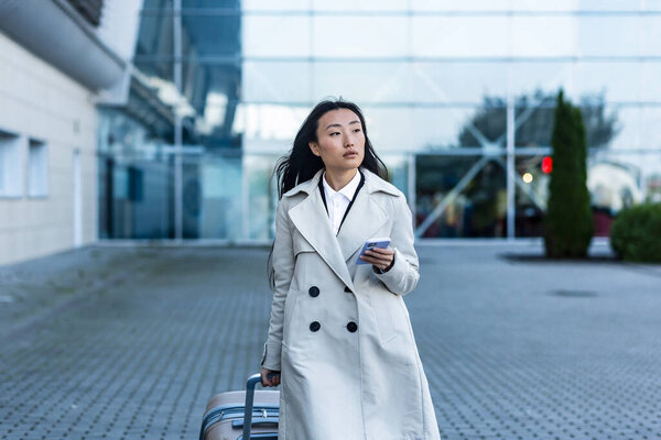 Beautiful female tourist near the airport, a Chinese woman walking with a big suitcase, an Asian woman holding a phone, using the application for booking accommodation and ordering a taxi
