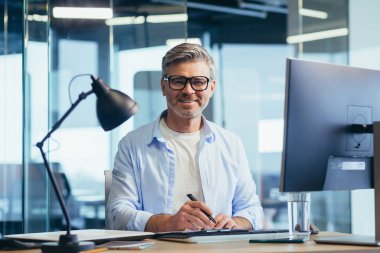 Gözlüklü başarılı kıdemli patron, bilgisayarda çalışıyor, iş adamı gömlekli, modern ofiste çalışıyor.