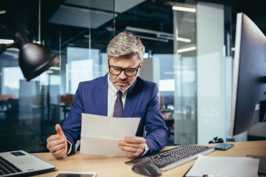 Experienced gray-haired businessman working at a computer, paperwork in the office, reading a letter from the bank