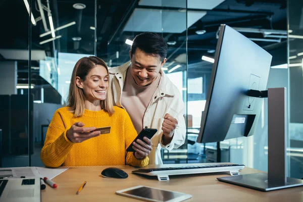 Asyalı erkek ve kadından oluşan yaratıcı bir ekip online alışveriş yapıyor, masada birlikte oturuyor, bir hediye seçiyor, neşeli ve gülümsüyor, akıllı telefon ve kredi kartı kullanıyor.