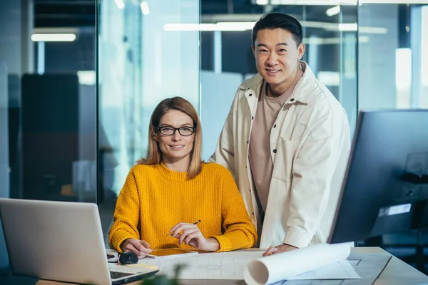 Modern bir ofisteki bir masada, Asyalı bir adam ve kadın kameraya bakıyor ve gülümsüyor, kağıt üzerinde bina planı yapıyor.