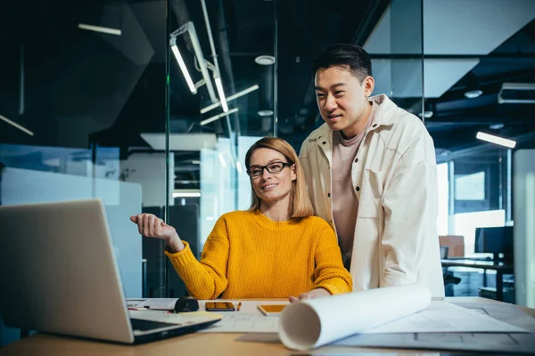 Tasarımcı ve mimarların iki çalışanı, projeyi düşünüyorlar, Asyalı bir adam ve sarışın bir kadın, gündelik giysiler içinde masada birlikte çalışıyorlar, bir plan çizimi projesiyle birlikte.