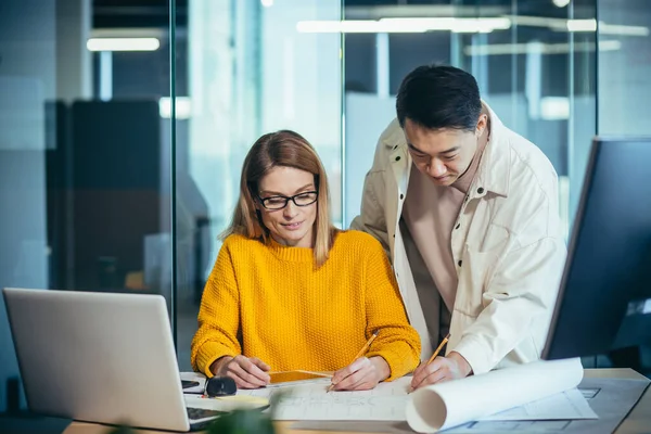 Tasarımcı ve mimarların iki çalışanı, projeyi düşünüyorlar, Asyalı bir adam ve sarışın bir kadın, gündelik giysiler içinde masada birlikte çalışıyorlar, bir plan çizimi projesiyle birlikte.