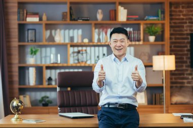Asyalı patronun elleri havada kameraya bakıyor ve gülümsüyor. Ofiste çalışan adam, başarılı ve olumlu iş adamı portresi.
