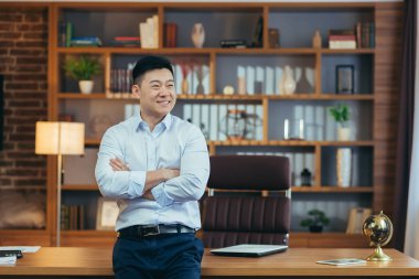 Klasik bir ofiste çalışan başarılı bir işadamının portresi, gömlekli Asyalı patron kollarını kavuşturup pencereye bakarken gülümsüyor ve seviniyor.