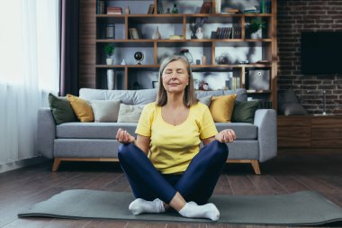 Yaşlı, kır saçlı kadın bir paspasın üzerinde meditasyon yapıyor, evde dinleniyor, yoga yapıyor.