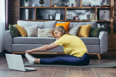Yaşlı gri saçlı kadın evde spor yapıyor, paspasın üzerinde oturuyor, çevrimiçi aerobik dersleri alıyor, esneme hareketleri yapıyor, dizüstü bilgisayara bakıyor.