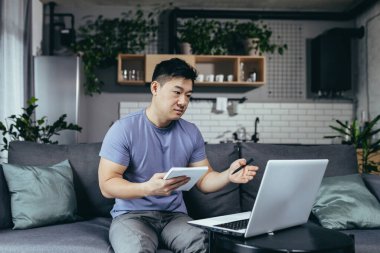 Evde okuyan bir adam, online öğrenim için dizüstü bilgisayar kullanıyor, bir Asyalı kanepeye oturuyor, yeni bilgiler yazıyor.