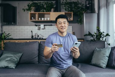 İnternetten mutlu Asya alışverişleri, evdeki kanepede oturan, elinde telefon ve banka kartı tutan adam.