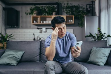 Telefondan kötü haberler okuyan bir adam, Asyalı hayal kırıklığına uğramış ve evde kanepede oturan telefon ekranına bakıyor.