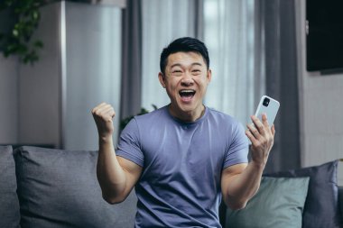 Evde mutlu bir adam, başarılı bir galibiyet, online olarak koltukta oturarak kazanılmış, Asyalı çığlıklar neşeyle ve gülümsemeyle kameraya bakıyor, telefonu tutuyor.