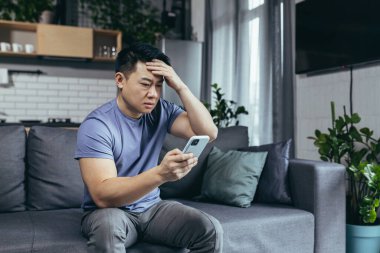 Endişeli adam internetten haberleri okuyor, telefon ekranına bakıyor, Asyalı koltukta oturuyor, ciddi ve üzgün.