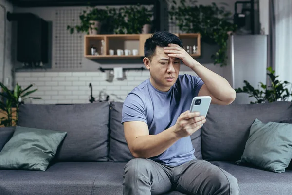 Telefondan kötü haberler okuyan bir adam, Asyalı hayal kırıklığına uğramış ve evde kanepede oturan telefon ekranına bakıyor.