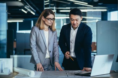 Çok ırklı iş mimarları ve tasarımcılarından oluşan bir ekip dizüstü bilgisayarın ekranına bakarak bir proje taslağı çizimi oluşturmayı tartışıyor. Asyalı erkek ve kadın müteahhitler takım çalışması.