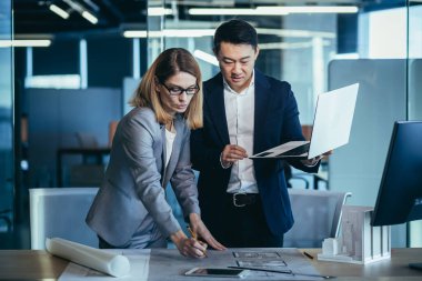 Çok ırklı iş mimarları ve tasarımcılarından oluşan bir ekip dizüstü bilgisayarın ekranına bakarak bir proje taslağı çizimi oluşturmayı tartışıyor. Asyalı erkek ve kadın müteahhitler takım çalışması.