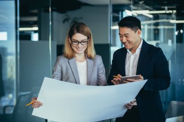 Grup genç yaratıcı mühendisleri modern ofiste yeni projeyi tartışıyorlar bir plan okuyorlar. Takım düşünceli mimar tasarımcısı. Dijital tablet kullanan Asyalı erkek ve kadın meslektaşlar