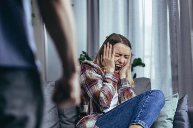 Aile içi şiddet, kızgın koca bağırıyor ve kadına vuruyor, yakın plan fotoğraf, karısı kanepede otururken ağlıyor.