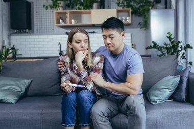 Asyalı erkek karısını destekliyor, hayal kırıklığı yaratan hamilelik testi sonuçları, koltukta oturan çok ırklı genç çift, hayal kırıklığına uğramış.