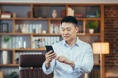 Klasik ev ofisinde çalışan Asyalı bir adamın yakın plan portresi, akıllı telefon ekranına bakan ve gülümseyen bir iş adamı.