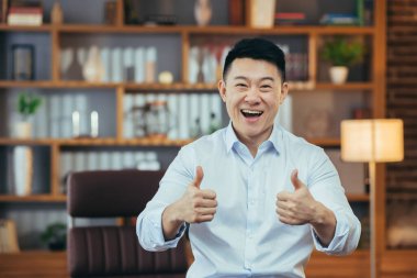 Klasik ofiste geç saatlere kadar çalışan başarılı Asyalı adam, kameraya bakıp baş parmağını gösteren mutlu iş adamı portresi.