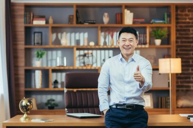 Asyalı patronun elleri havada kameraya bakıyor ve gülümsüyor. Ofiste çalışan adam, başarılı ve olumlu iş adamı portresi.