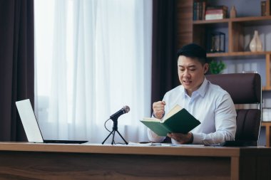 Asyalı vaiz internetten İncil okur, bir podcast kaydeder, masaya oturur ve kameraya bakar, mikrofon kullanır, elini kalbinde tutar, çevrimiçi radyo yayını yapar.