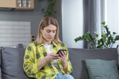 Ciddi ve odaklanmış bir kadın evinde, koltukta oturmuş, düşünceli bir şekilde telefondan haberleri okuyor.
