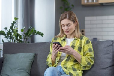 Evde mutlu bir kadın telefonla kitap okuyor ve gülümsüyor, mutfakta kanepede oturuyor.