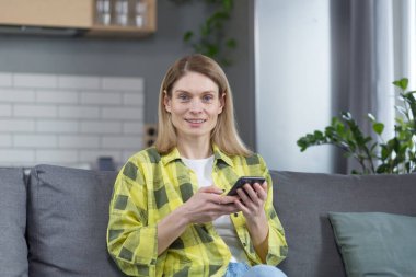 Mutlu orta yaşlı sarışın kadın evinde koltukta oturuyor kameraya bakıyor ve elinde telefonla gülümsüyor.