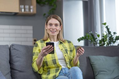 Evdeki mutlu bir kadının portresi, kanepeye oturmuş mutlu bir şekilde kameraya bakıyor gülümsüyor, telefonu tutuyor ve online alışveriş için banka kartı tutuyor.