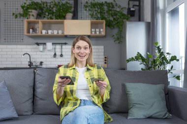 Evdeki mutlu bir kadının portresi, kanepeye oturmuş mutlu bir şekilde kameraya bakıyor gülümsüyor, telefonu tutuyor ve online alışveriş için banka kartı tutuyor.