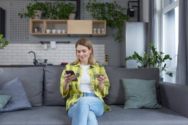 İnternetten alışveriş yapan mutlu bir kadın. Kanepede oturuyor, telefon ve banka kartı kullanıyor.