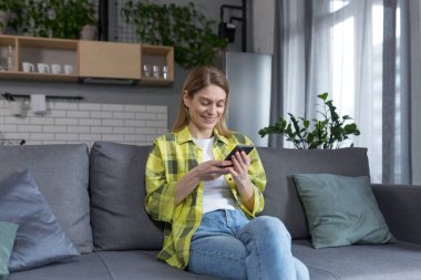 Evde mutlu bir kadın telefonla kitap okuyor ve gülümsüyor, mutfakta kanepede oturuyor.