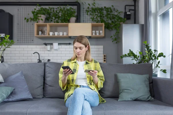 Odaklanmış kadın internetten alışveriş yapmayı seçiyor, ciddi eş elinde telefon ve banka kartıyla koltukta oturuyor.
