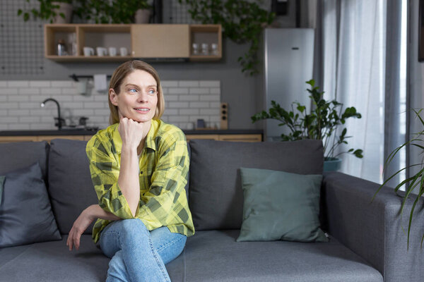 Женщина среднего возраста сидит дома на диване и думает: