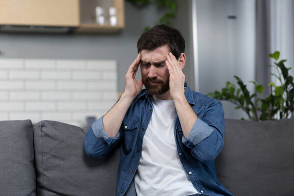 Мужчина болен дома, у него сильная головная боль, сидит на диване на кухне