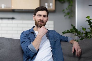 Evde sakallı bir adamın portresi, koltukta oturuyor ve kameraya bakıyor.