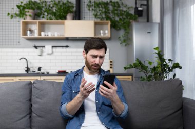 Adam evde, şok olmuş ve hayal kırıklığına uğramış, telefona bakıyor, kanepede oturuyor.