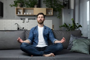 Evde tek başına sakallı genç bir adam, kanepede oturuyor ve nilüfer pozisyonunda meditasyon yapıyor, günlük kıyafetlerle.