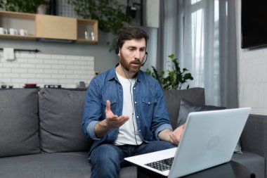 Adam evden uzaktan çalışıyor. Bilgisayarı ve kulaklığı internet üzerinden arama yapmak için kullanıyor.