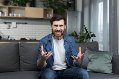Sinirli bir adam video görüşmesinde konuşuyor, web kamerasına bakıyor, öfkeyle el kol hareketi yapıyor, evde kanepede oturuyor.